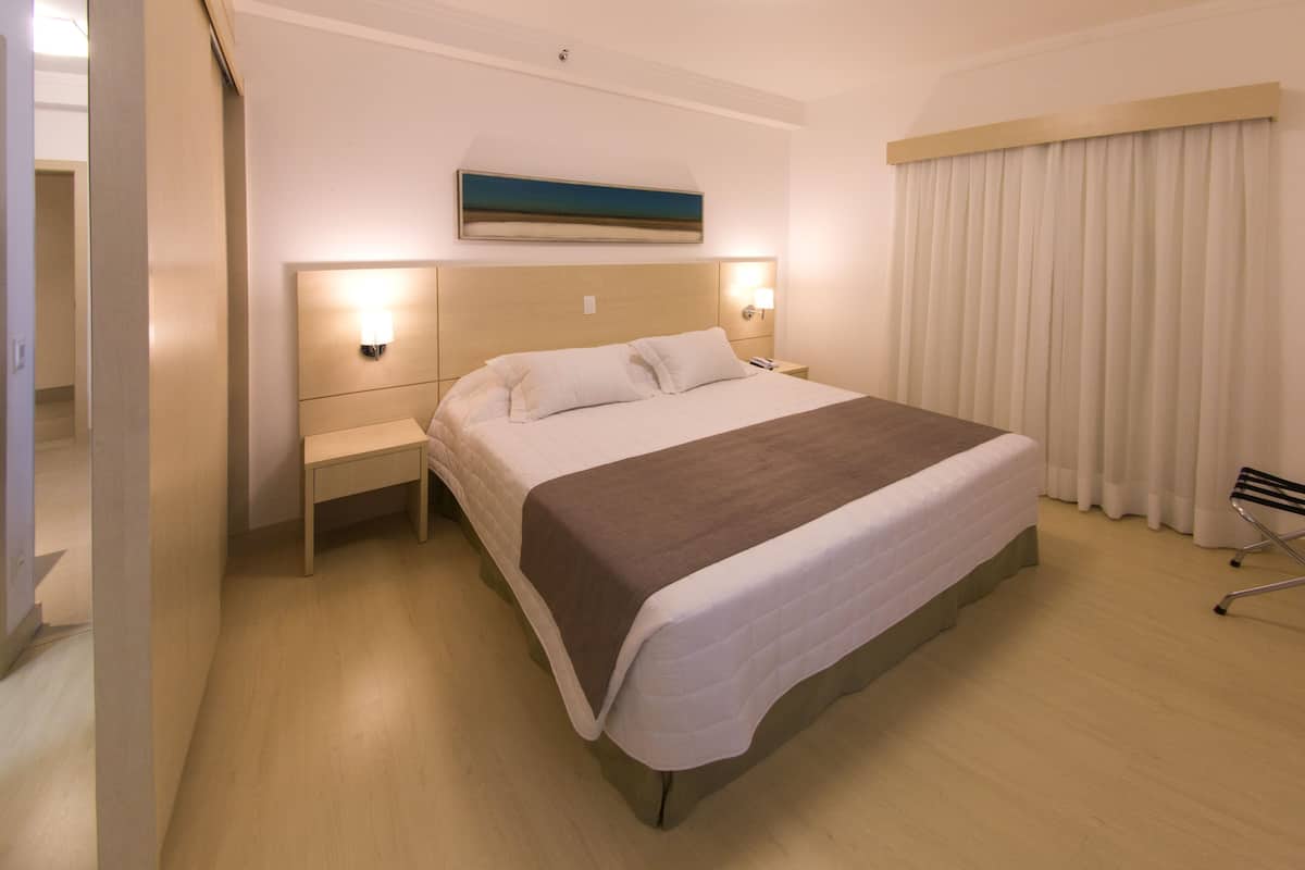 Une chambre d’hôtel avec un grand lit, des tables de chevet, une petite table, une lampe fixée au mur et une fenêtre avec des rideaux.