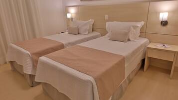 Een hotelkamer met twee bedden, elk met een beige sprei en witte kussens. Er zijn twee nachtkastjes, waarvan er één een lamp en notitieboekje heeft. De kamer heeft een houten vloer en een schilderij aan de muur.