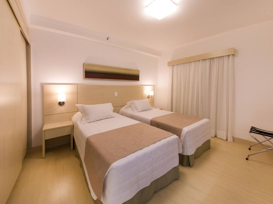 Quarto de hotel com duas camas, uma mesinha de cabeceira, uma cadeira e um quadro na parede.
