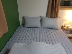 Ein ordentlich bezogenes Bett mit einer gesteppten, grauen Bettdecke und zwei Kissen, ein Nachttisch aus Holz mit einer Lampe und einer kleinen Portion Essen, sowie eine grüne Wand.