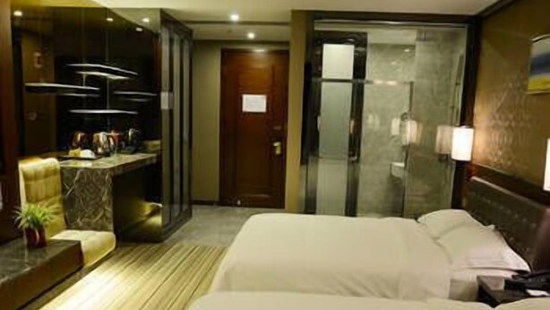 A hotel room with two beds, a sitting area with a chair and a small table, a minibar, and a glass-enclosed shower area. - Enping Jinzhou Holiday Hotel (Jiangmen)
