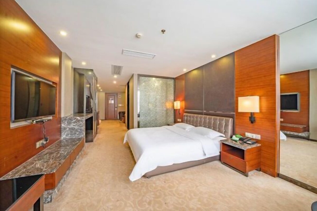 A hotel room with a large bed, a TV, a desk with a lamp, and a mirror.