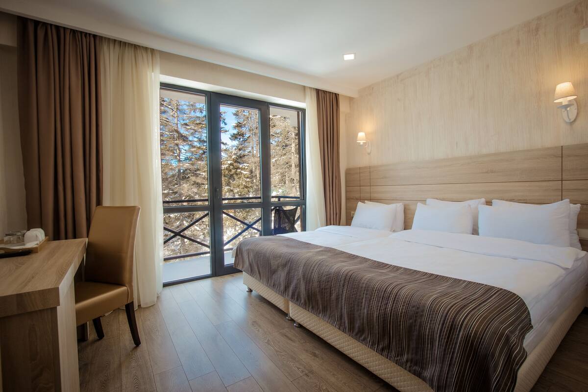 A hotel room with a large bed, a desk, and a view of trees through a sliding door.
