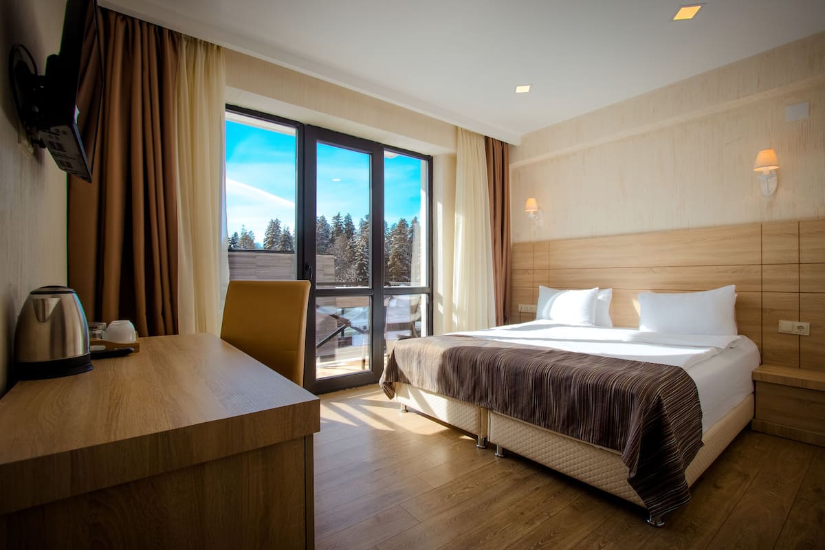 A hotel room with a bed, a desk, a chair, a coffee maker, and a view of trees through a large window.