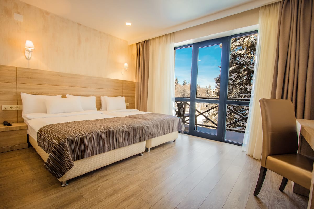 A modern hotel room with a large bed, a wooden headboard, a view of trees through a large window, and a seating area with a table and chair.