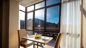 A dining area with a table set for two, a bottle of wine, and a bowl of olives. Large windows offer a view of a mountainous landscape. - BM Plaza (Borjomi)