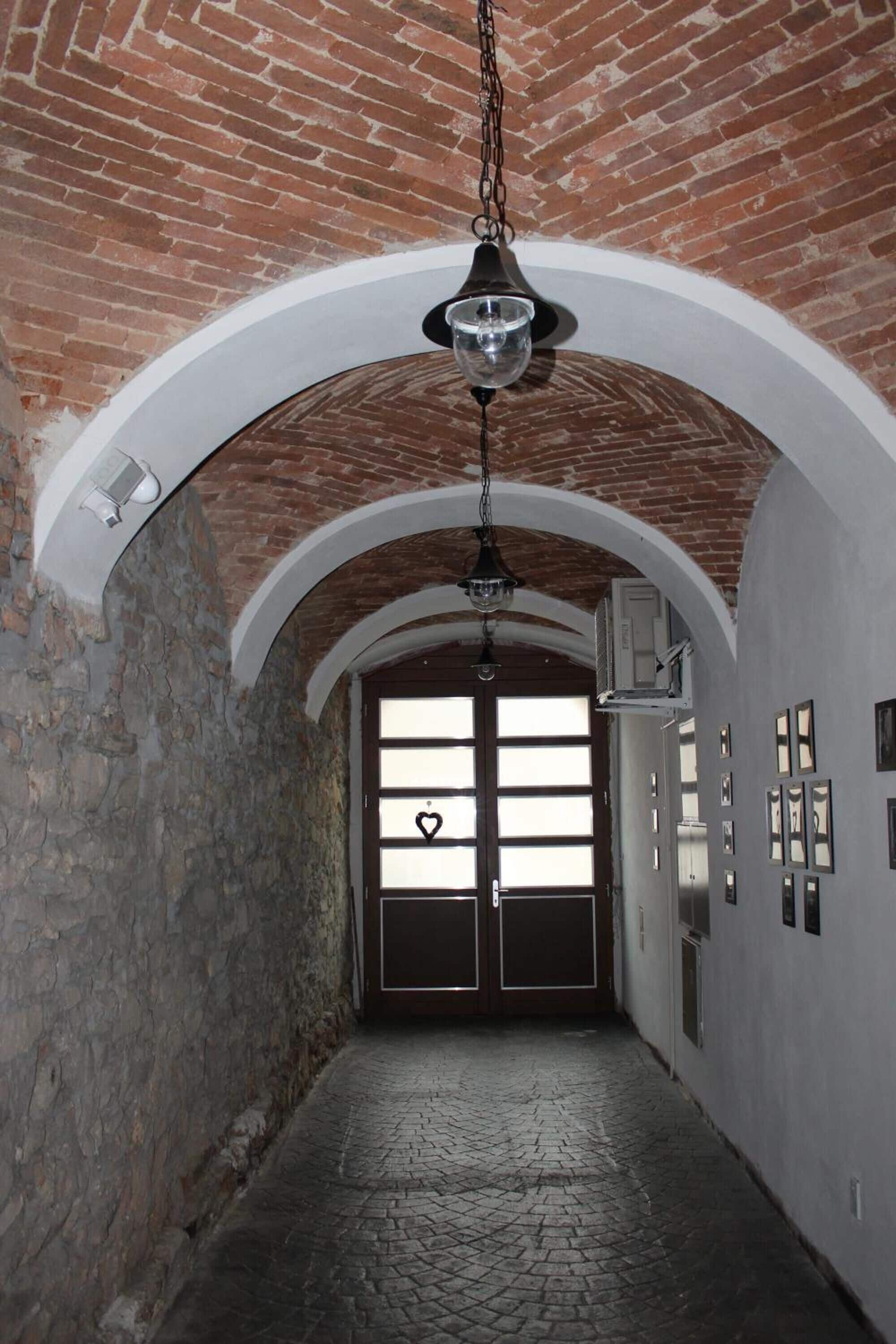 Un couloir avec un plafond en briques, des arches blanches, un luminaire suspendu et une porte ornée d’un symbole en forme de cœur.