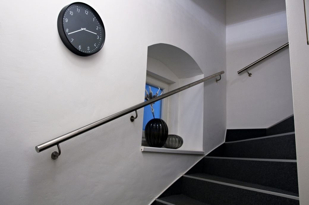 Una escalera con barandilla metálica, un reloj negro en la pared y un ventanal pequeño con marco azul.