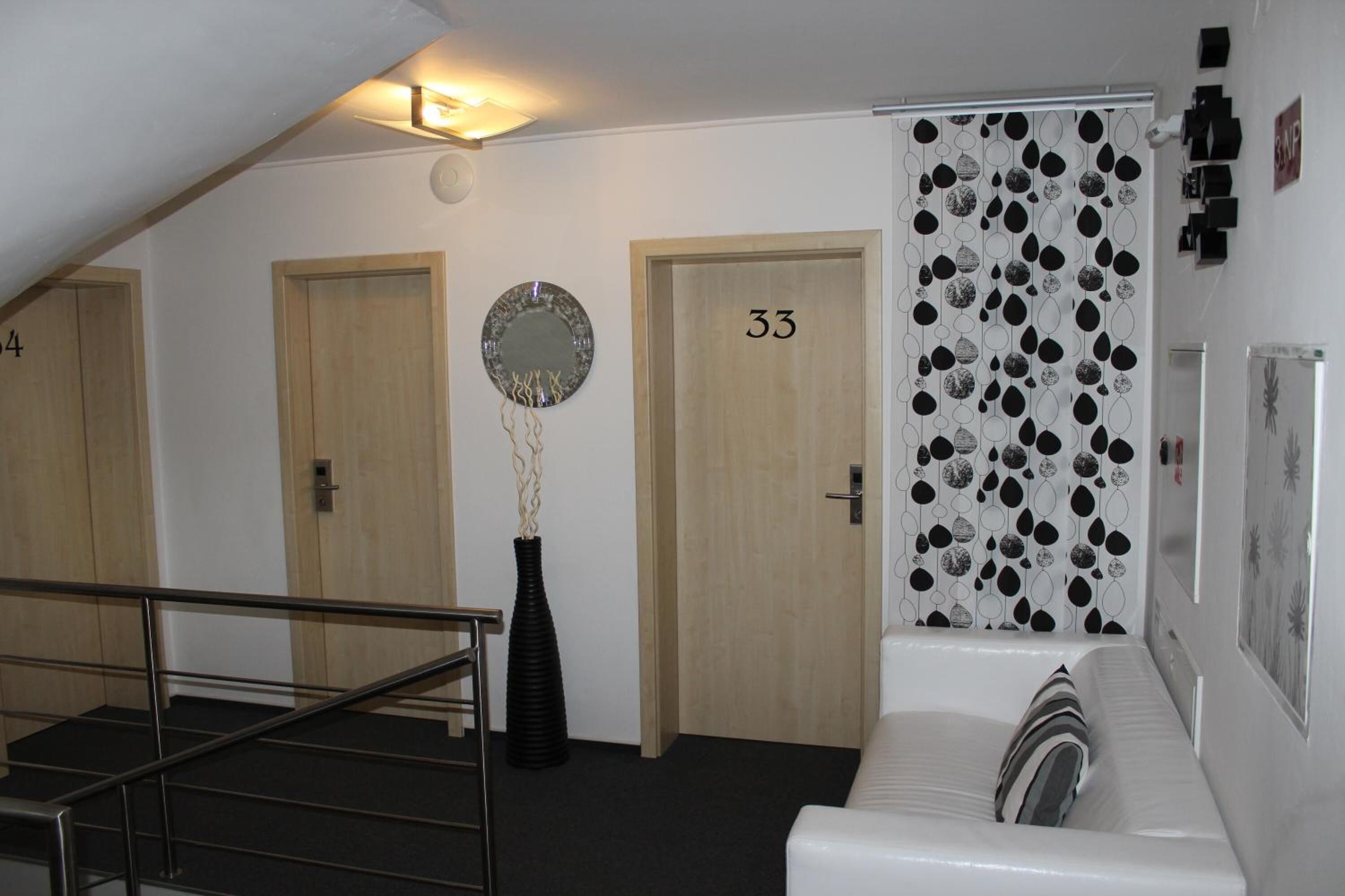 Un hall d’hôtel avec un escalier, trois portes numérotées 34, 35 et 33, un vase noir et un canapé blanc avec un coussin.