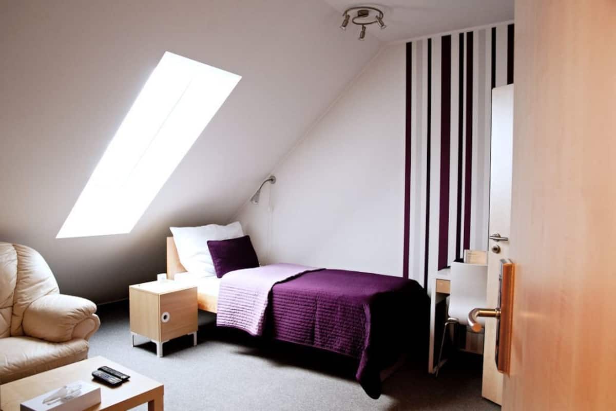 A bedroom with a sloped ceiling, a bed with a purple blanket, a nightstand, a chair, a desk, and a wardrobe.