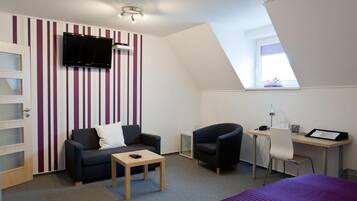 A modern room with a sloped ceiling, a TV mounted on the wall, a desk with a chair, a sofa, and a bed.