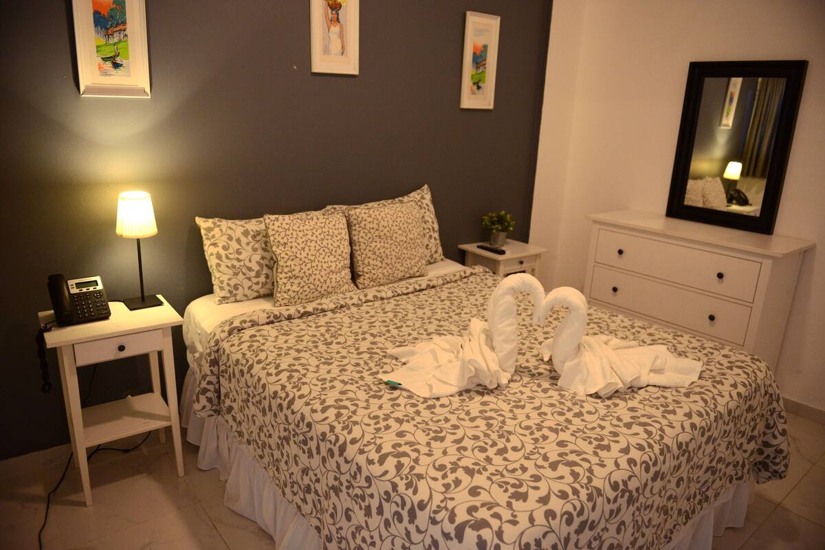 Una cama bien hecha con una colcha estampada, una lámpara de noche, una mesita con un teléfono y un espejo en la cómoda.
