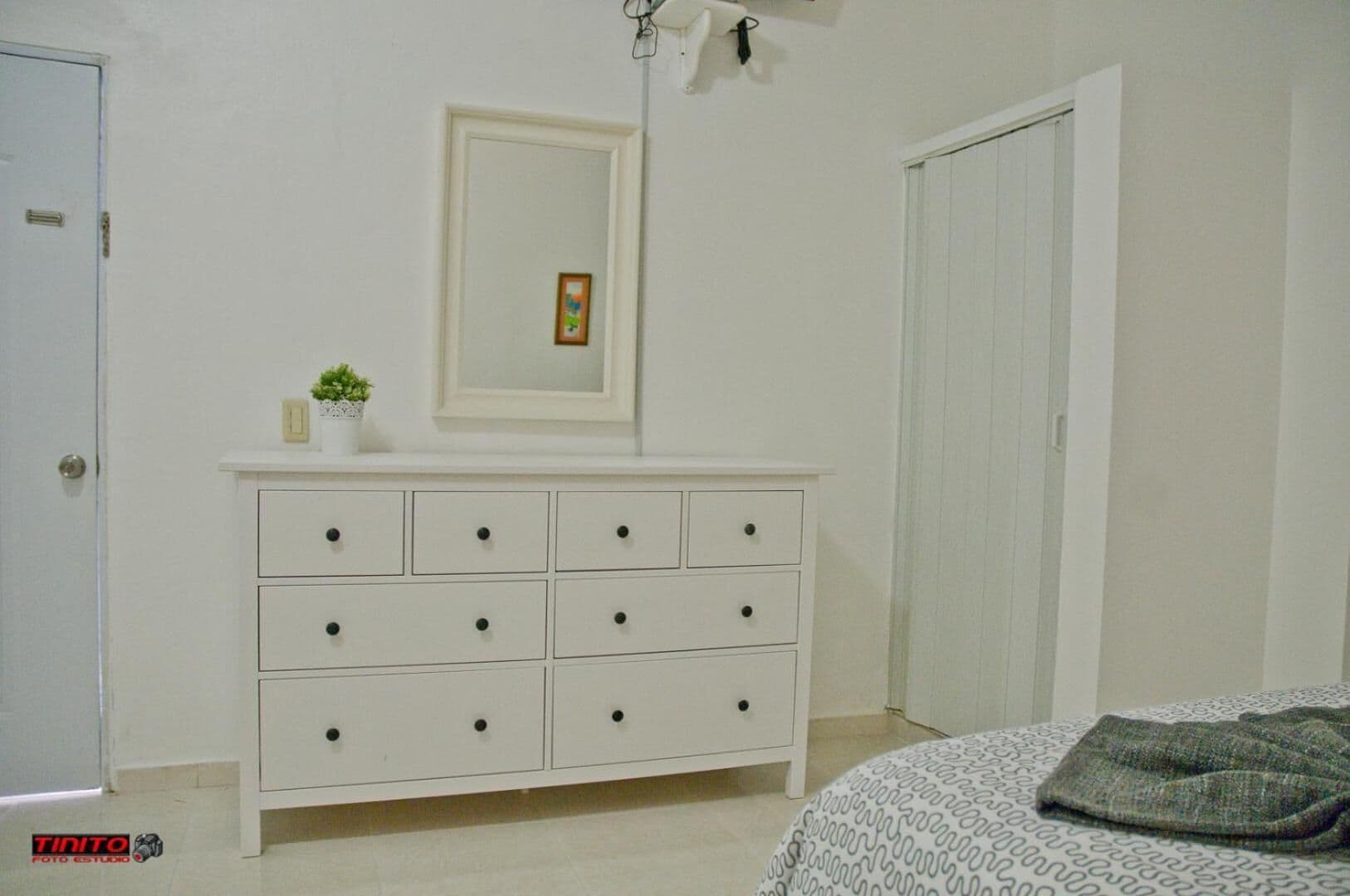 Un dormitorio con un aparador blanco, una cama con ropa de cama estampada, un cuadro en la pared y una puerta.