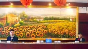 A reception desk with a large painting of a sunflower field, two individuals seated behind it, and red hanging decorations. - Hanting Hotel (Shaoxing)