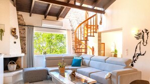 A modern living room with a stone wall, a wooden staircase, a grey sofa, a glass coffee table, and a fireplace. - Villa Claire Corfu (Corfu)