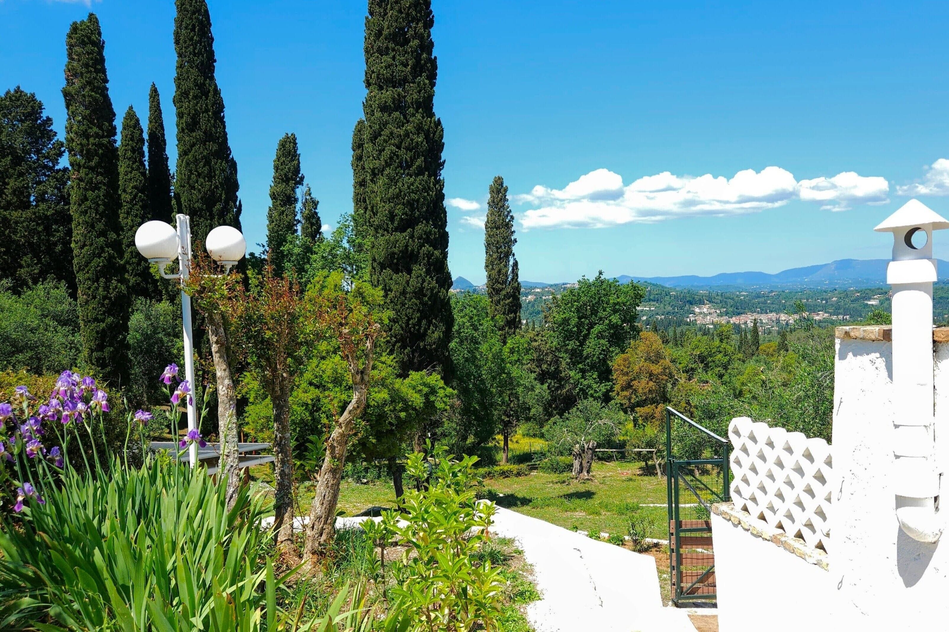 A pathway leading to a white building with a tower, surrounded by greenery and tall trees, overlooking a distant landscape.