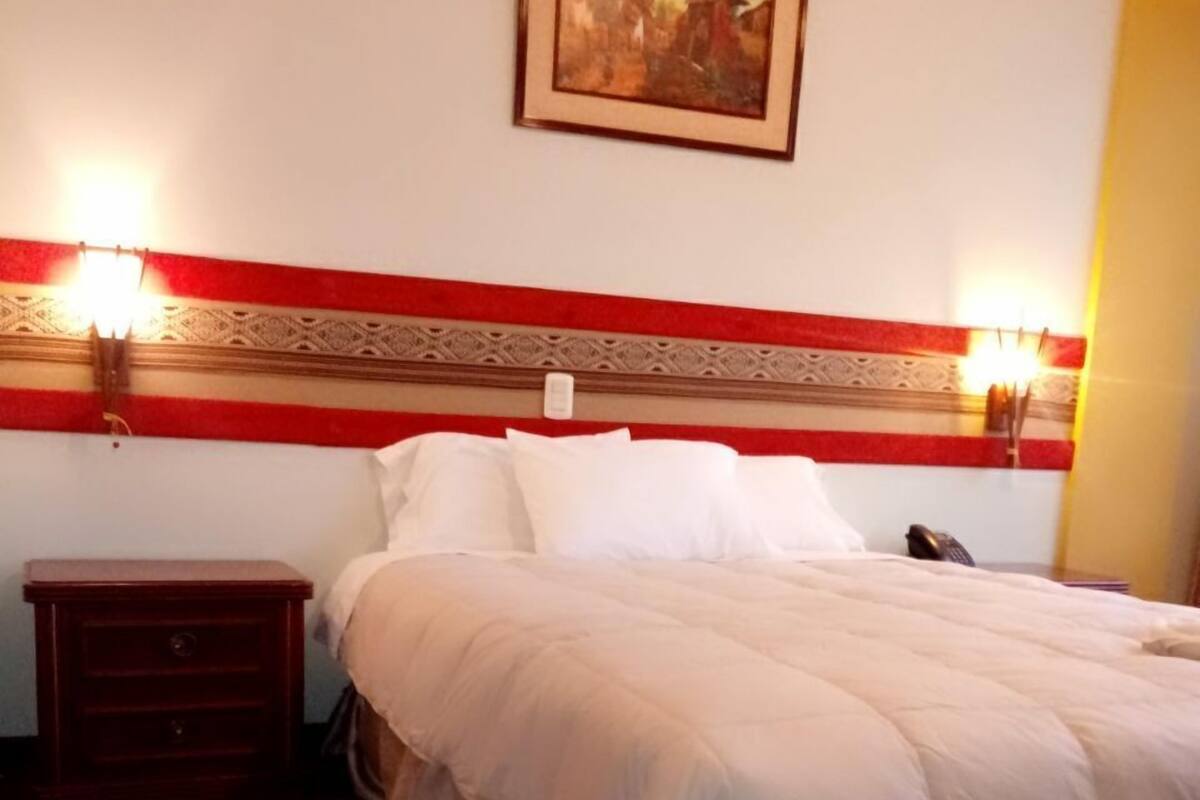 A neatly made bed with a white comforter and pillows, a wooden nightstand with a lamp, and a framed picture on the wall.