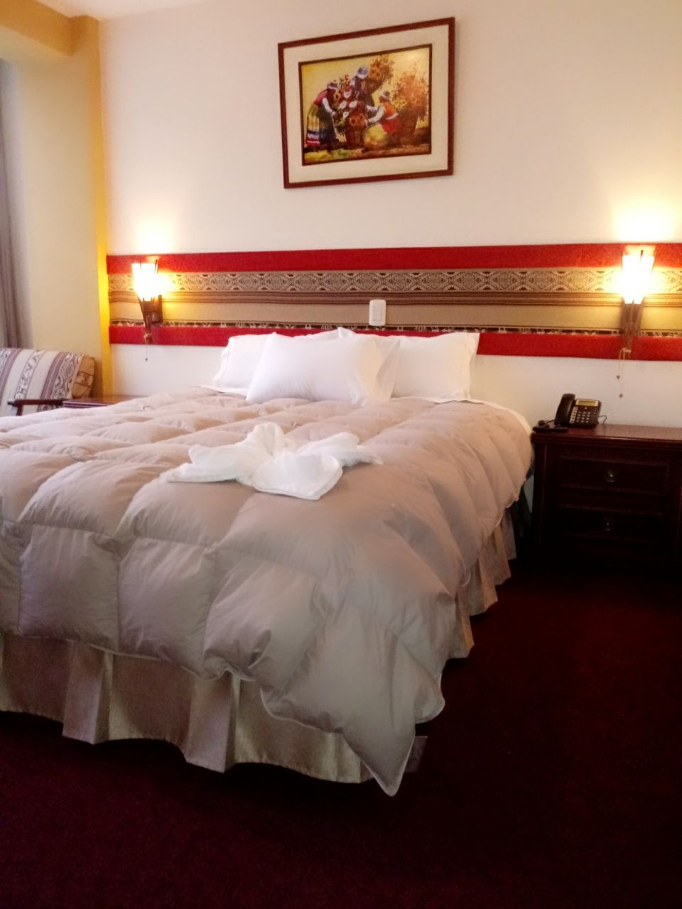 A neatly made bed with a red and white headboard, a bedside table with a lamp, and a painting on the wall.