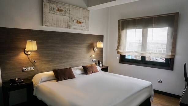 A hotel room with a large bed, two bedside lamps, a nightstand with a phone, a painting on the wall, and a window with sheer curtains. - Suites A Coruña (A Coruña)