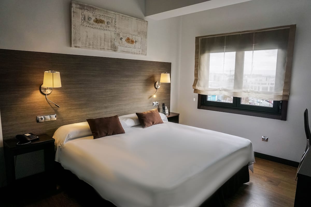 A hotel room with a large bed, two bedside lamps, a nightstand with a phone, a painting on the wall, and a window with sheer curtains.