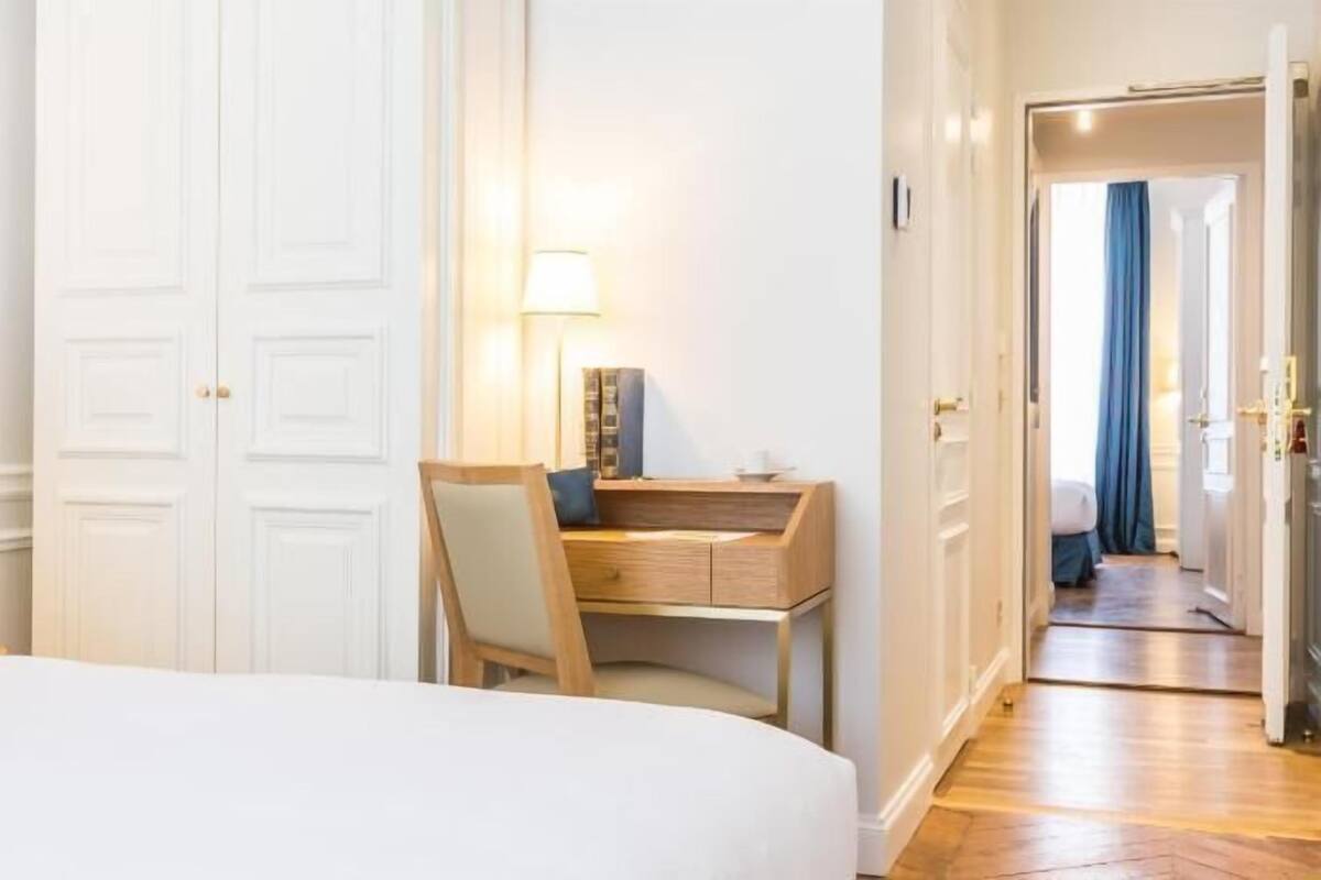 A hotel room with a bed, a desk with a chair, a lamp, and a door leading to another room.