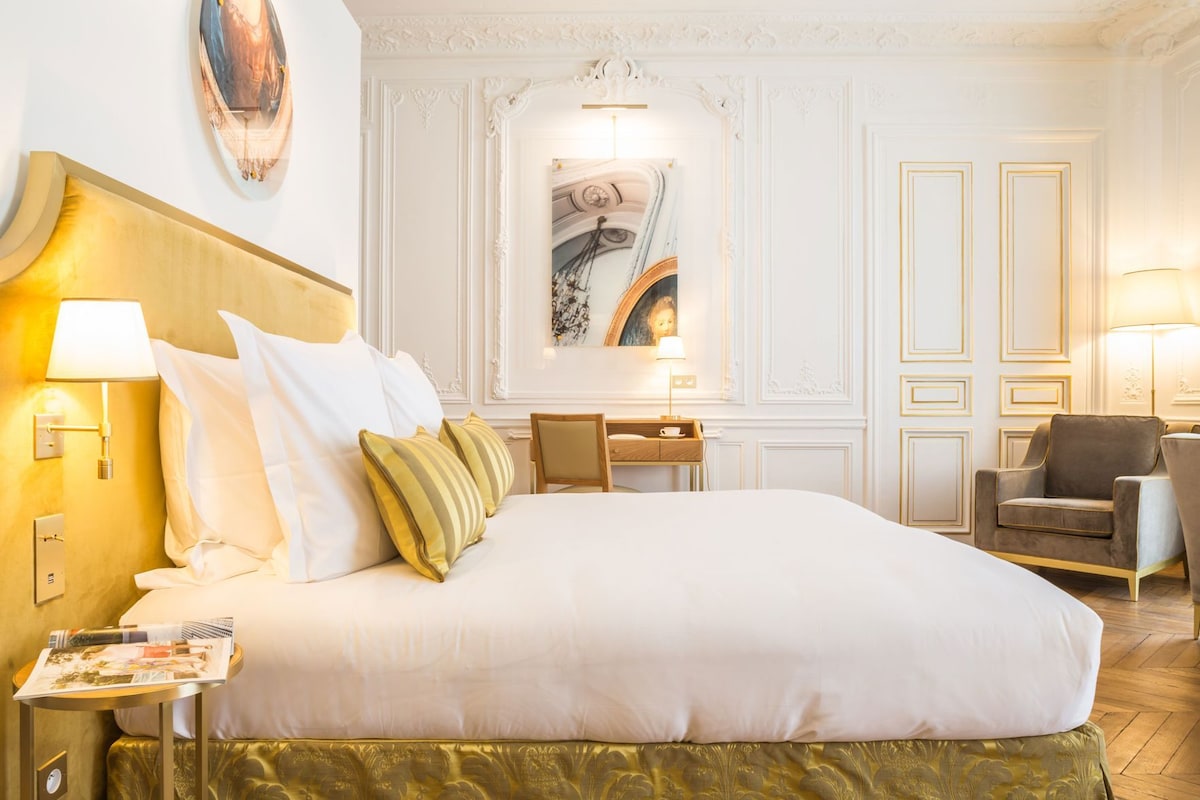 A hotel room with a large bed, a desk, a chair, and a wall-mounted painting.