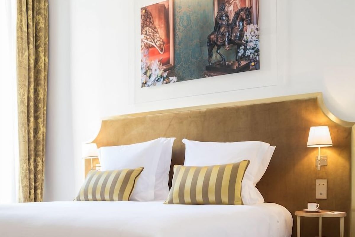 A hotel room with a large bed, two bedside lamps, a framed artwork above the bed, and a small table with a cup.