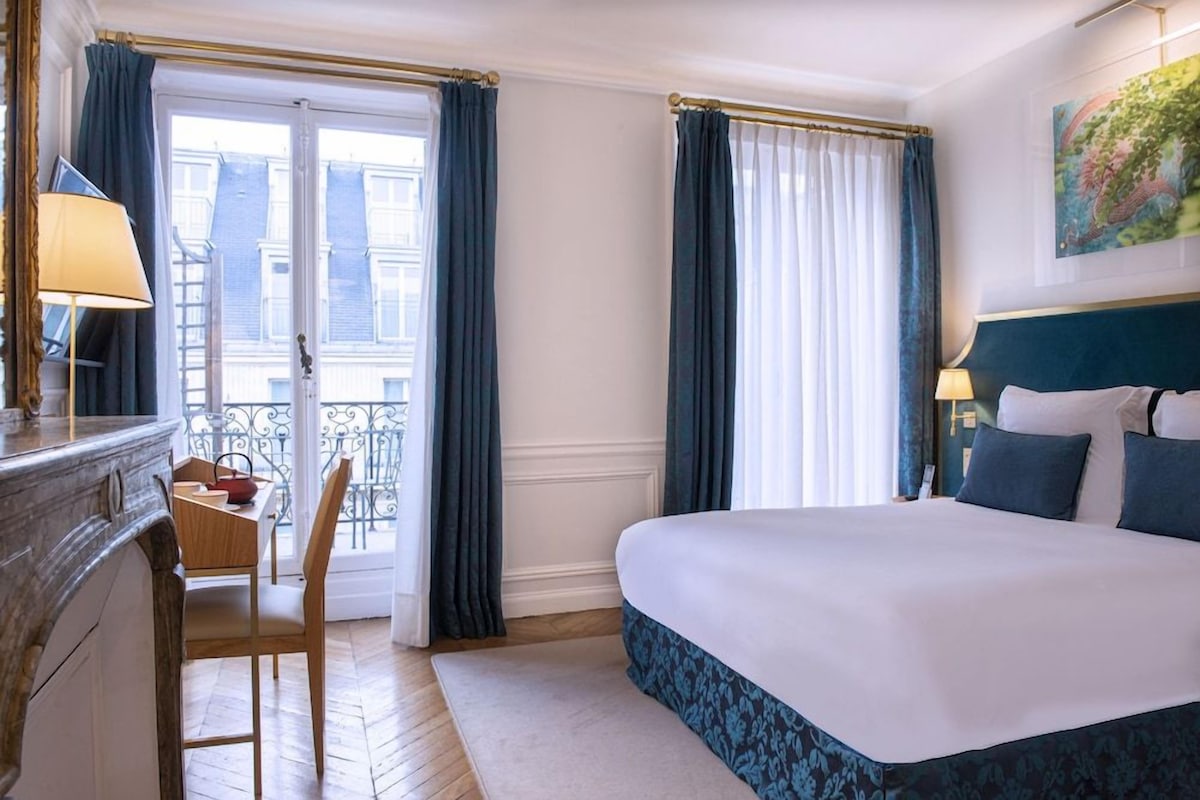 A hotel room with a bed, a desk with a chair, a window with curtains, and a painting on the wall.