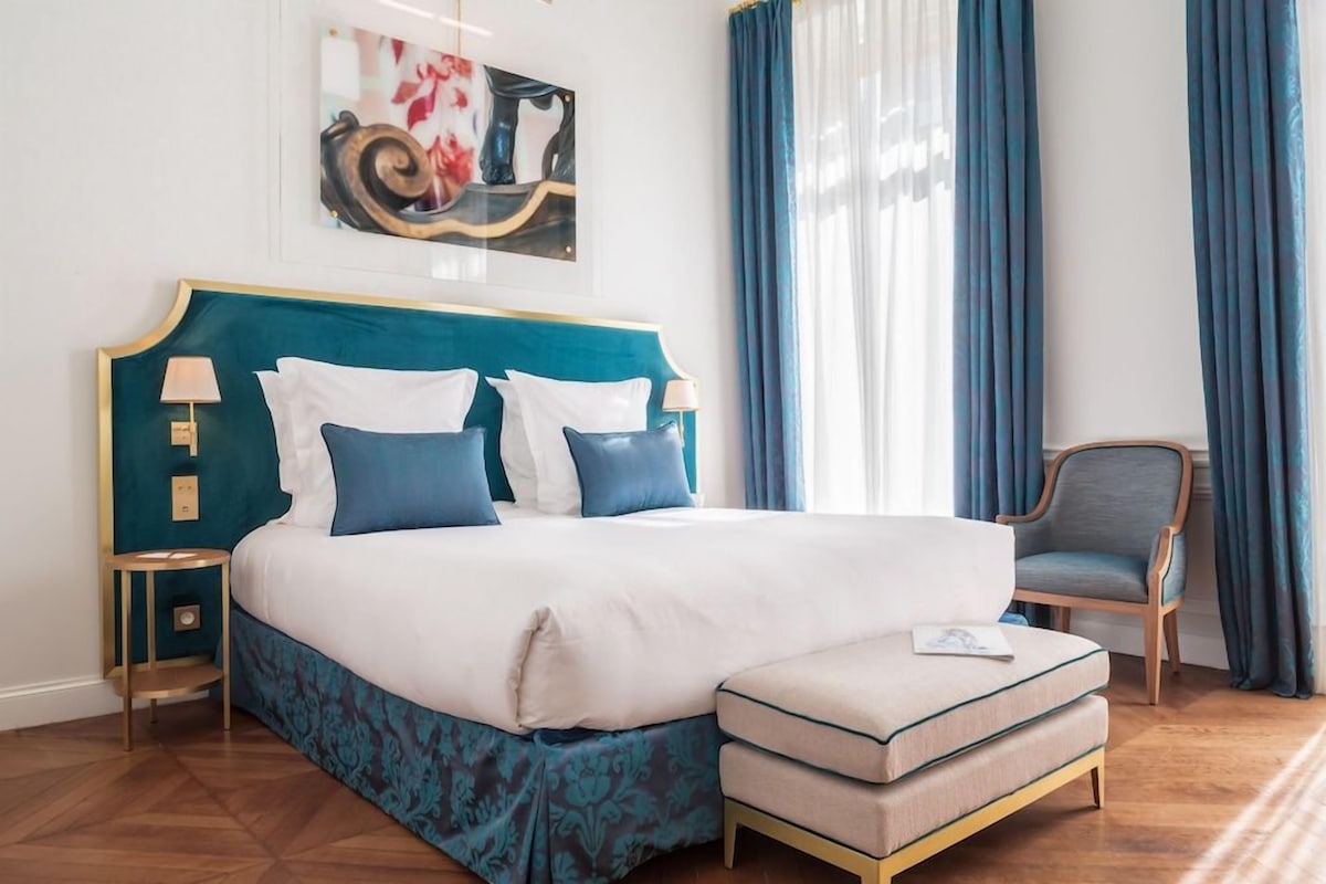 A neatly arranged bedroom with a large bed, bedside tables, a chair, and a painting on the wall.