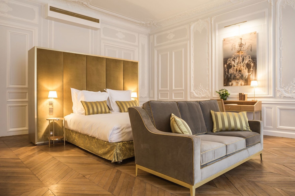 A hotel room with a large bed, a sofa, a nightstand, a lamp, and a painting on the wall.