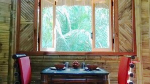 Interior de madeira com uma mesa e cadeiras, uma janela com vista para vegetação e um telhado de palha.