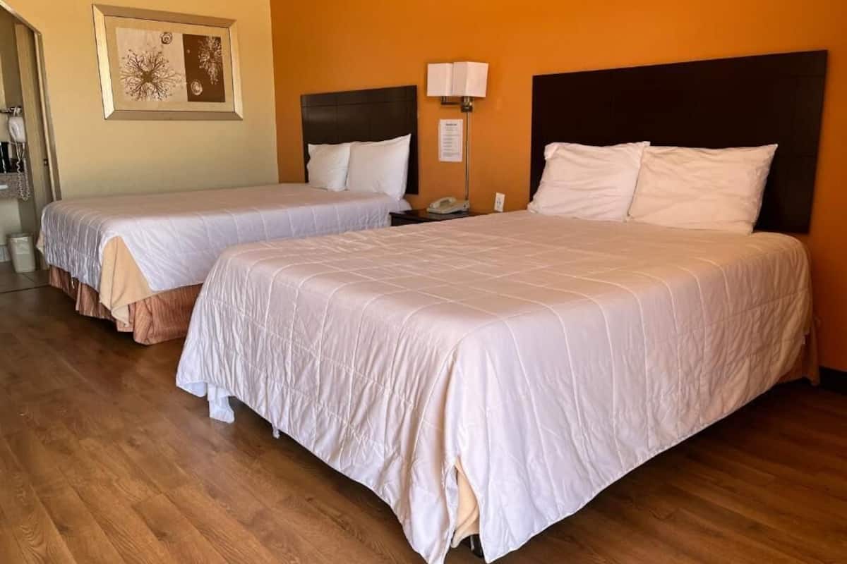 A hotel room with two beds, wooden flooring, a nightstand with a lamp, and a framed picture on the wall.