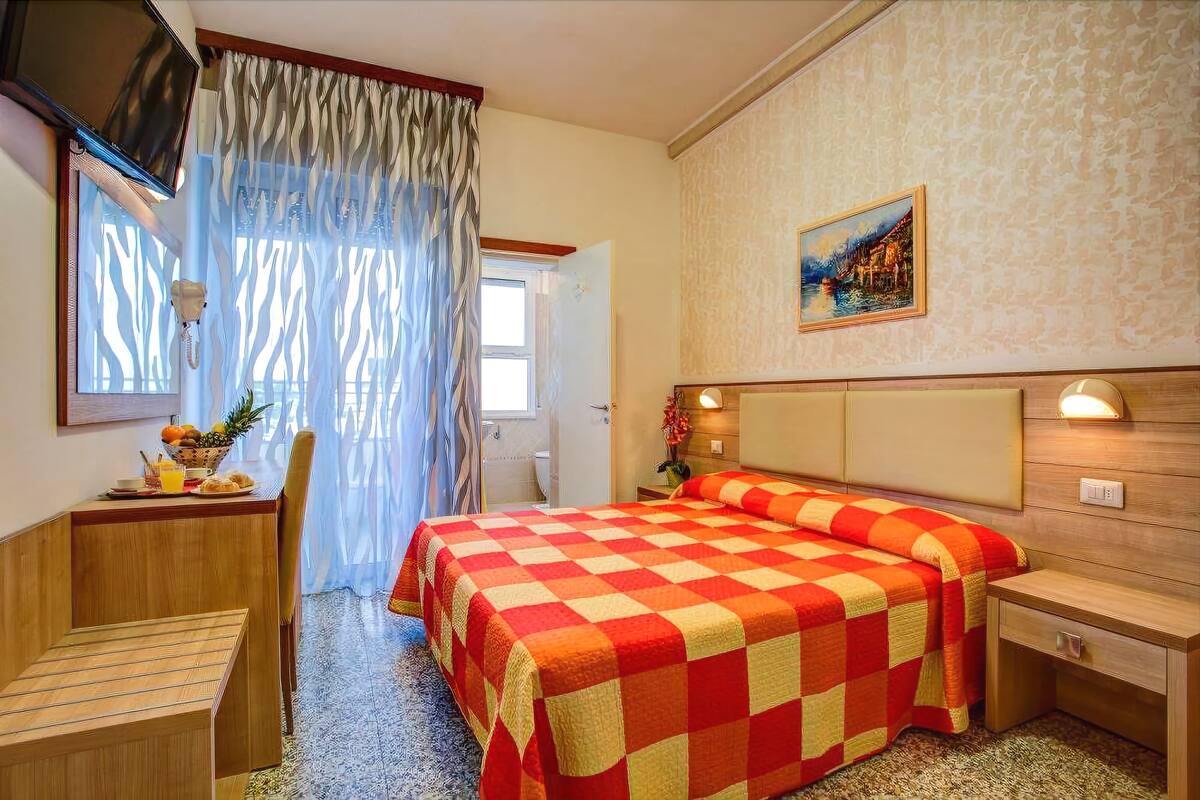 A hotel room with a checkered bedspread, a desk with a chair, a television, and a painting on the wall.