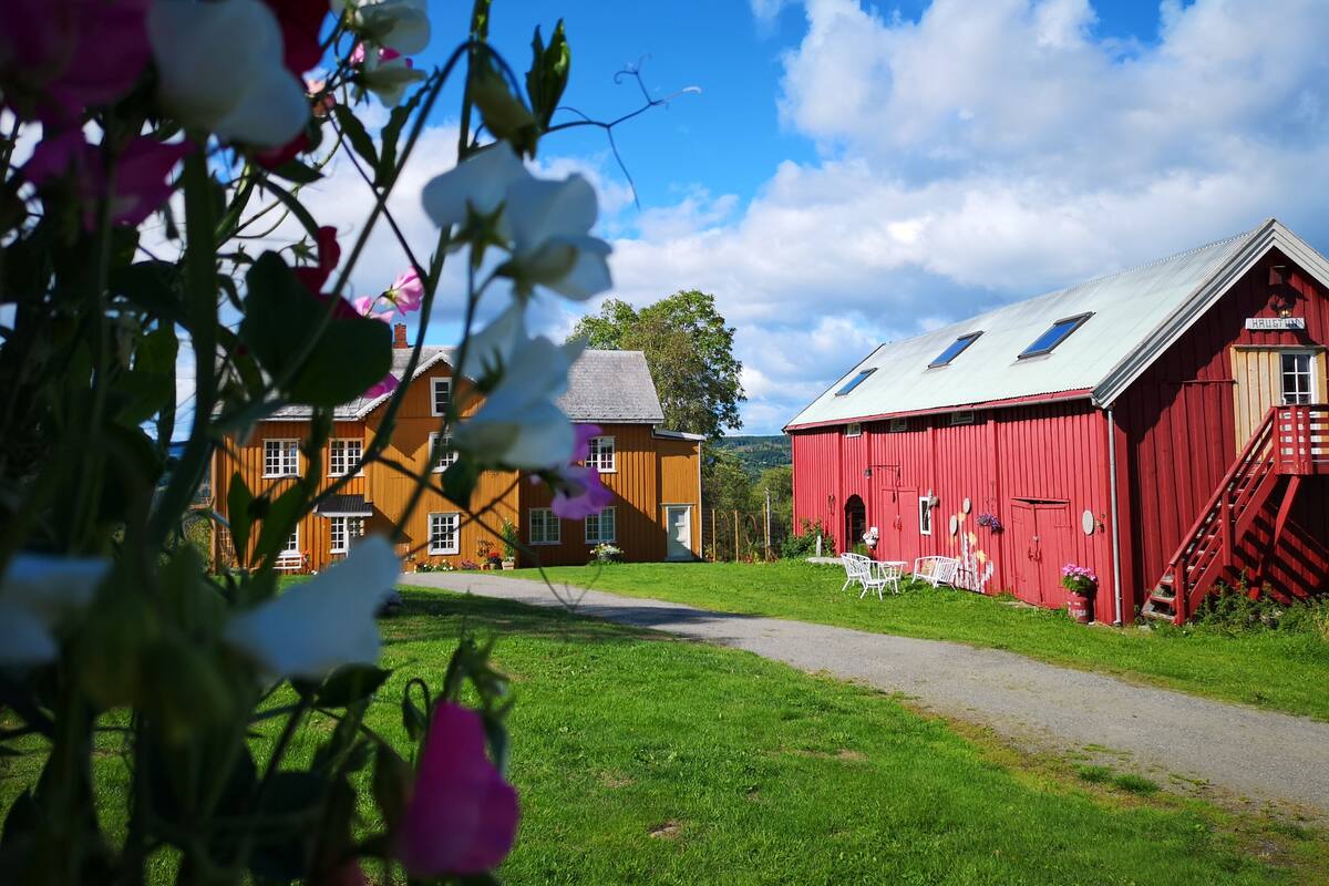 Ein rotes Bauernhaus mit Metalldach, ein Holzhaus mit grauem Dach und ein klarer, blauer Himmel.