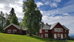 A red log cabin with a white door and a smaller adjacent cabin, surrounded by lush green grass and tall trees. - Haugtun gårdspensjonat og kulturverksted (Nordre Land)