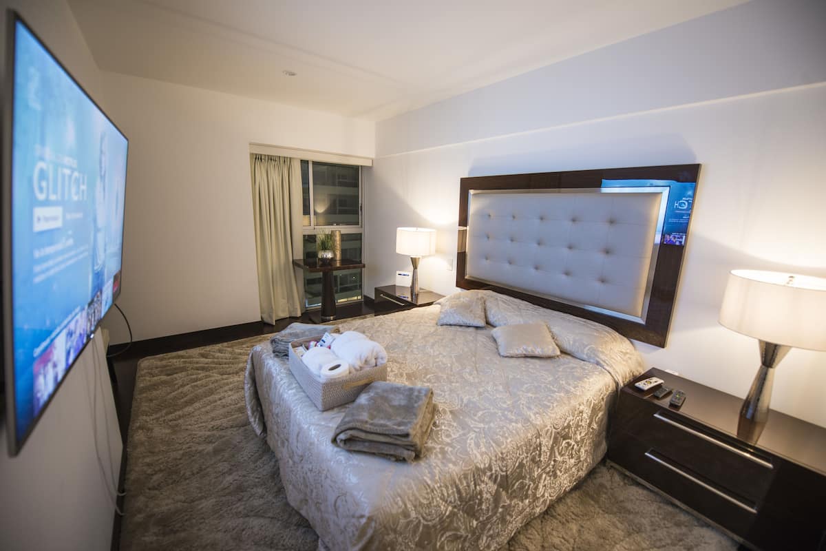 A hotel room with a large bed, a flat-screen TV mounted on the wall, a bedside table with a lamp, and a window with curtains.