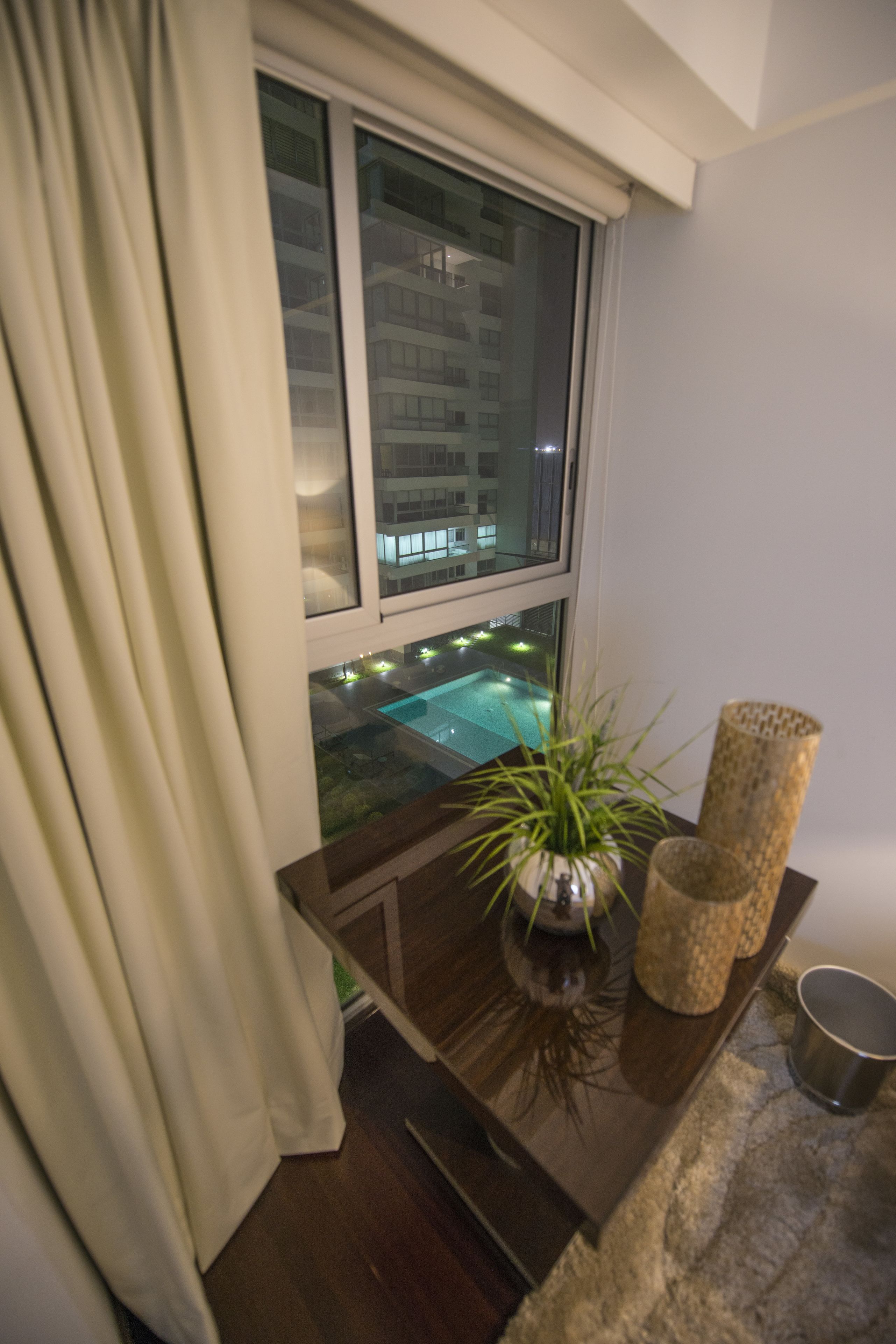 Habitación con ventana que da a una piscina, una planta en maceta y una pequeña mesa con una vela y una taza.