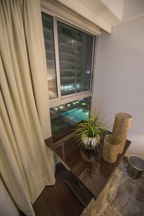 A room with a window overlooking a pool, a potted plant, and a small table with a candle and a cup. - Moon Luxury Apartments (Santiago de Surco)