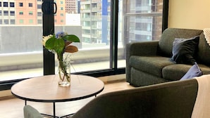 A modern living room with a round table, a green armchair, and a large window offering a city view.