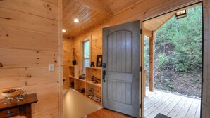 Interior de uma cabana de madeira com uma porta de madeira que dá para um deck. Prateleiras são visíveis no interior da cabana, e uma pequena mesa com uma tigela está à esquerda.
