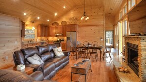 Interior acolhedor de uma cabana, com sofá de couro, mesa de jantar, cadeiras, cozinha e lareira.