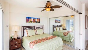 Une chambre à coucher comprenant un lit, une table de chevet, une lampe, un ventilateur de plafond et un grand miroir.