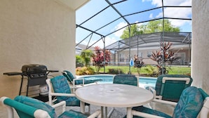 Eine Terrasse im Freien mit weißen Korbmöbeln und einem runden Tisch mit Blick auf einen Swimmingpool und einen angelegten Garten.