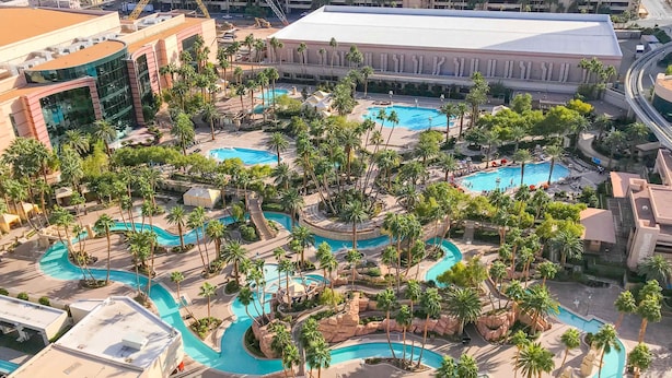 Vista aérea de um resort com várias piscinas, palmeiras e um edifício grande ao fundo.