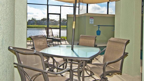 Un patio esterno con tavolo in vetro e sedie in vimini, un ombrellone beige e vista su un corpo d'acqua.