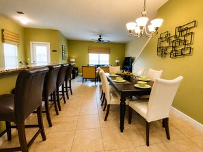 A dining area with a long table, chairs, and a chandelier. There is a kitchen area with a counter and chairs.
