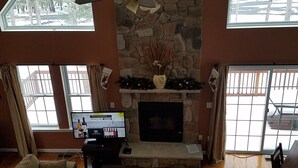 A spacious living room with a stone fireplace, a ceiling fan, and large windows offering a view of the outdoors. - Mountain/lake community in Poconos,PA (Lackawaxen)
