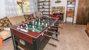 A room with a foosball table, a sofa, a chair, and a wooden shelf with decorative items.