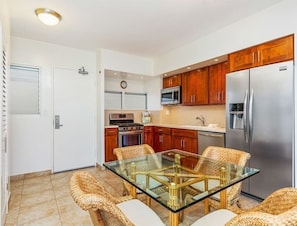 A modern kitchen with wooden cabinets, stainless steel appliances, and a glass dining table with wicker chairs. - Touch the Palms! Island-Style Condo w/Free WiFi, Full Kitchen–Waikīkī Shore #316 (HONOLULU)
