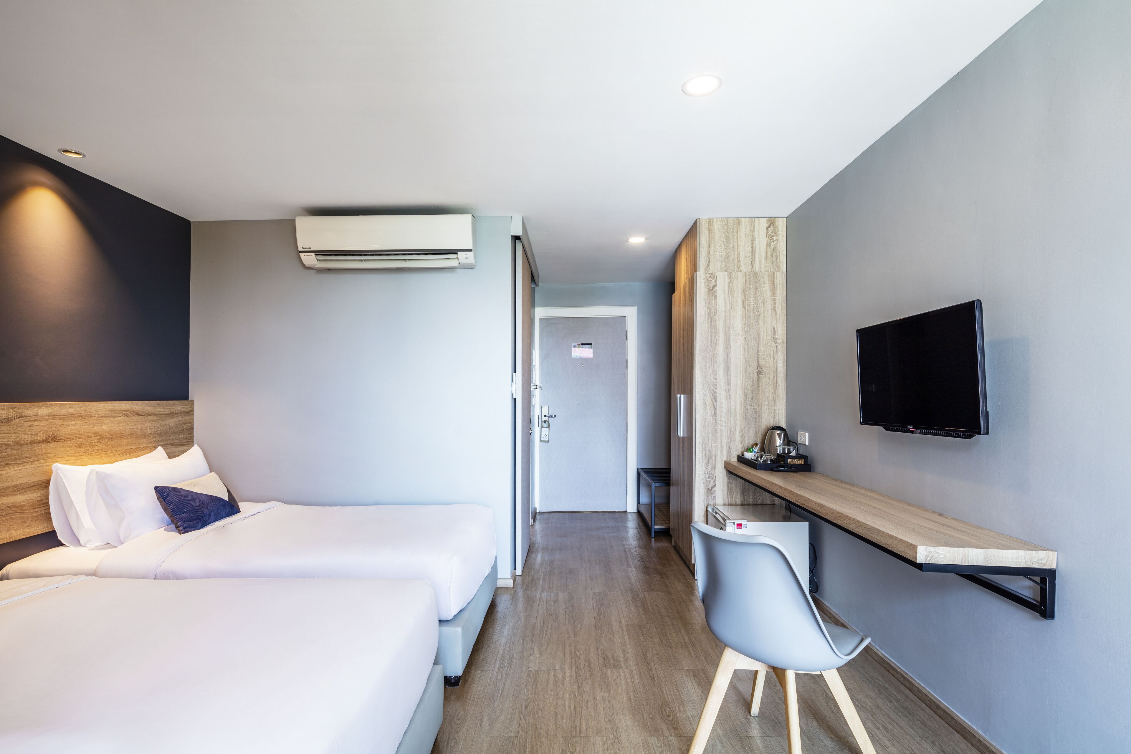 A modern hotel room with a bed, bedside table, chair, TV, and an air conditioning unit.